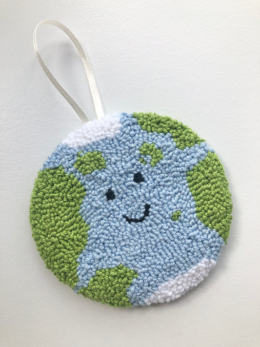 A hand-punched cotton yarn wall hanging depicting a smiling Earth with blue oceans and green landmasses, backed with felt and equipped with a ribbon for hanging.